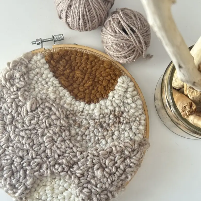 Textured fiber art hoop in cream and mustard beside balls of cotton yarn and driftwood