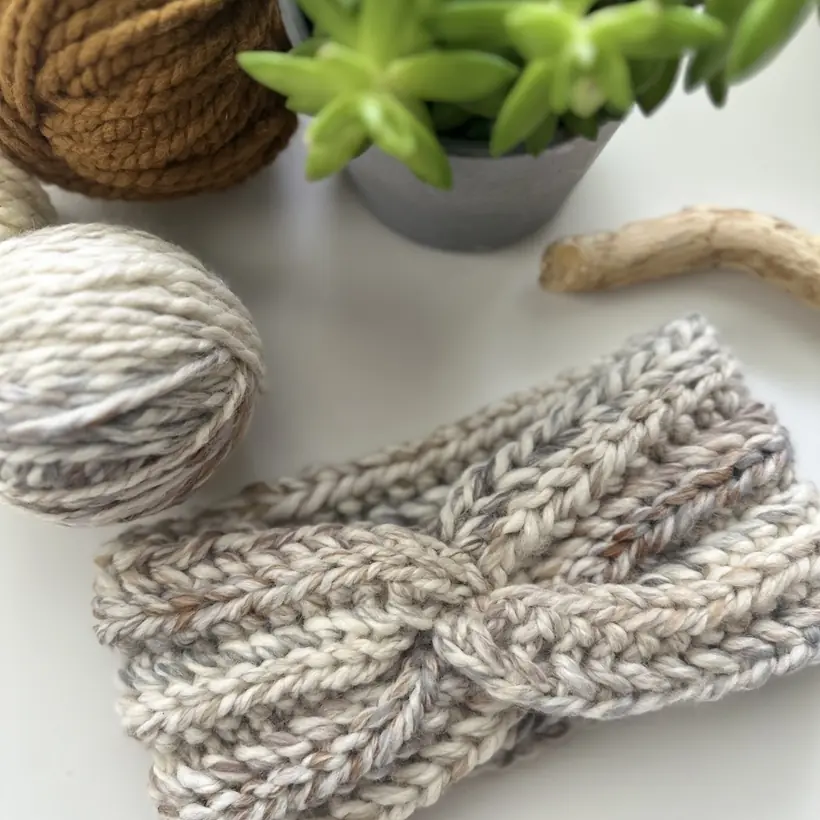 Hand-knit headband in cream and grey beside balls of natural cotton yarn